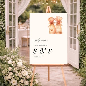 Op de afbeelding: Een crèmekleurig welkomstbord voor een bruiloft op een houten ezel. Het bord toont het woord "welcome" in cursief, de namen "S & F" en de datum 23.09.2024. Twee aquarel illustraties van cadeauzakjes staan bovenaan.