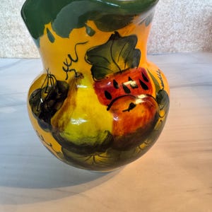 May include: A ceramic pitcher with a bright yellow base colour, featuring hand-painted illustrations of various fruits including a pear and a red fruit with black seeds. The rim is painted in a deep green, and the surface has a glossy finish.