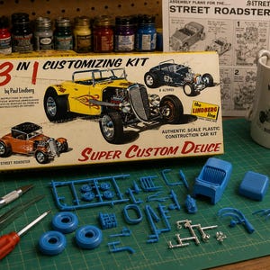 May include: A 3-in-1 Customising Kit by Paul Lindberg for a Super Custom Deuce model car. The box features illustrations of three car variations. Included are blue plastic model parts, a hobby knife, a screwdriver, and assembly instructions.