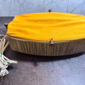 May include: An electric warming tray with a rectangular shape, featuring a textured tan and brown woven-look exterior. The top surface is covered in a vibrant mustard yellow fabric, secured by small brown knobs at the centre and sides.