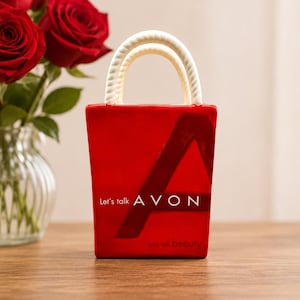 Pode incluir: Uma bolsa de cerâmica vermelha com alças brancas e o logotipo da Avon. A bolsa tem o texto "Let's talk AVON" e "let's talk beauty" impresso. Um buquê de rosas vermelhas está ao fundo.