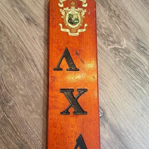 Könnte beinhalten: Ein langes, rechteckiges Holzpaddel mit orangefarbenem Beizfinish. Es zeigt ein Wappen an der Oberseite, die griechischen Buchstaben Lambda Chi Alpha in Schwarz und den Text "WAYNE '52" in Schwarz am unteren Ende. Am Griff befindet sich ein Loch.