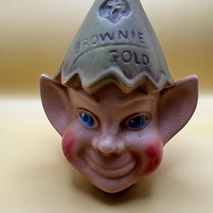 May include: A ceramic figurine head of a brownie elf with large pointed ears, bright blue eyes, and rosy cheeks. The elf wears a green pointed hat with the embossed text "BROWNIE GOLD" and a small clover symbol on the front of the hat.