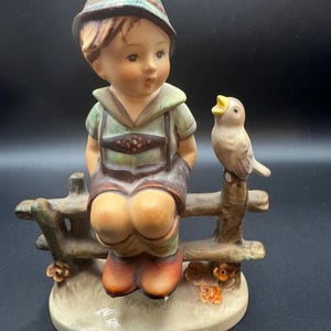 May include: A ceramic figurine of a young boy in traditional Bavarian attire, seated on a wooden fence. A small bird perches nearby. The figurine features detailed painting with a muted color palette, including green, brown, and orange.