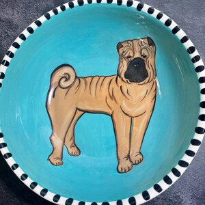 May include: A round ceramic bowl featuring a hand-painted tan Shar-Pei dog with characteristic skin folds and a black muzzle. The interior of the bowl is a vibrant turquoise blue, and the rim is decorated with a black and white dashed pattern.