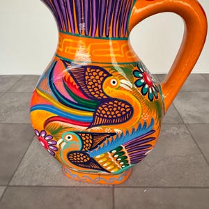 May include: A bright orange ceramic pitcher featuring hand-painted, colorful bird motifs in shades of blue, yellow, and purple. The vessel includes a curved handle, a wide spout, and intricate geometric patterns encircling the neck and base.