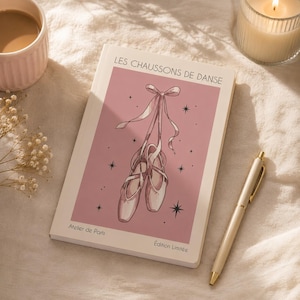 May include: A rectangular notebook with a white cover featuring a pink central panel, an illustration of ballet slippers with ribbons, and the text LES CHAUSSONS DE DANSE, Atelier de Paris, and Édition Limitée printed in black ink.