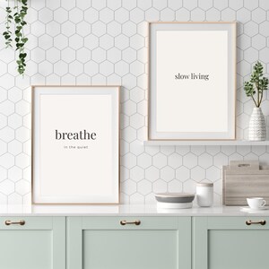 Puede incluir: Dos impresiones artísticas enmarcadas con marcos dorados. Una impresión dice "breathe in the quiet" y la otra "slow living". Las impresiones se muestran en una encimera blanca con un mueble verde menta.