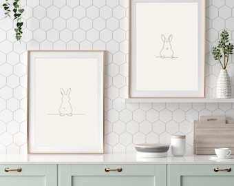 May include: Two framed art prints with minimalist line drawings of rabbits. The artwork is in a light beige colour, with a thin black outline of a rabbit. The frames are a light brown colour. The prints are displayed in a kitchen setting.