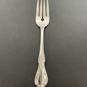 May include: A silver fork with four tines and a decorative handle. The tines are evenly spaced, and the handle features an ornate, symmetrical design. The fork is made of a shiny, reflective metal.
