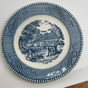 May include: A blue and white decorative plate with a detailed pastoral scene in the center. The plate has a textured blue border with a repeating pattern and decorative scrollwork. The rim is white with a dotted design.