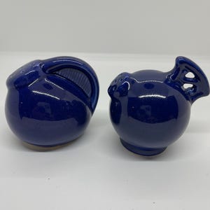 Puede incluir: Un par de saleros y pimenteros de cerámica azul cobalto. El salero tiene un asa y una abertura ranurada. El pimentero tiene un asa y varios agujeros pequeños. Ambos tienen un acabado brillante y una forma redondeada.