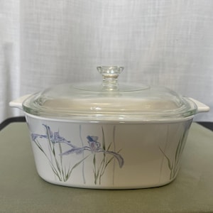 Pode incluir: Travessa branca com tampa de vidro transparente. A travessa apresenta um desenho floral de íris roxas e caules verdes. A tampa tem um botão de vidro transparente. A travessa tem duas pegas brancas em ambos os lados.