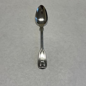 May include: A silver spoon with a shell design on the handle. The spoon has a rounded bowl and a long, slender handle. The spoon is made of a shiny, reflective metal.
