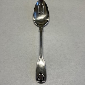 May include: A silver slotted spoon with an oval bowl and a long handle. The bowl has two curved slots. The handle has a decorative shell design at the end. The spoon is made of a shiny metal.