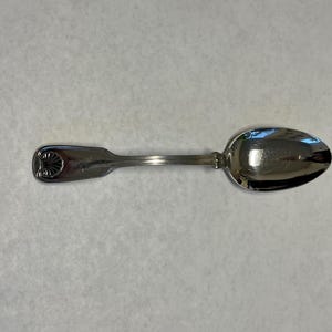 Puede incluir: Una cuchara de plata con un diseño de concha en el mango. La cuchara tiene un mango largo y delgado y un cuenco redondeado. La cuchara está hecha de un metal brillante y reflectante.