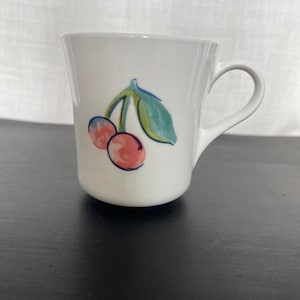 May include: White ceramic mug with a curved handle. The mug features a hand-painted design of two red cherries with a green leaf. The cherries have pink highlights. The mug is on a black surface.