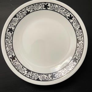 May include: White ceramic plate featuring a black floral and swirl pattern around the edge. The plate's design is simple and elegant, suitable for serving food. The black pattern contrasts with the white surface.