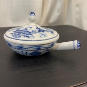 May include: A small, white porcelain serving dish with a lid, featuring a blue floral pattern. The dish has a handle and a decorative finial on the lid. The design is a classic blue and white pattern, suitable for serving sauces or dips.