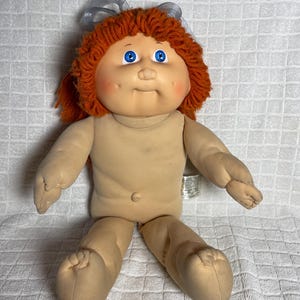 Puede incluir: Una muñeca con cuerpo de tela beige suave, cara redonda y ojos azules brillantes. La muñeca presenta cabello de lana naranja texturizado con un lazo de cinta gris translúcido en la parte superior. Tiene un pequeño ombligo y mejillas rosadas.