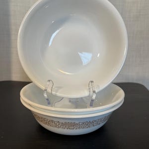 May include: Three white ceramic bowls, stacked. The top bowl is plain, the lower two feature a brown floral pattern around the rim. The bowls are displayed on a black surface.