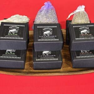 May include: An arrangement of black boxes with a bison logo, labeled "Sage", "Lavender", and "Cedar", alongside bundles of dried herbs and a wooden tray. The boxes are stacked on a wooden tray against a red background.