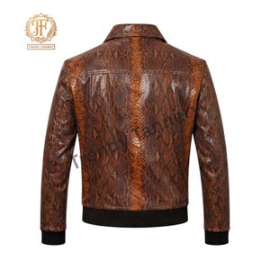 May include: A brown jacket featuring a snakeskin pattern throughout the exterior. The garment includes a pointed collar, long sleeves with black ribbed cuffs, and a black ribbed hem at the waist. The Trendy Tannery logo is visible in the top left corner.