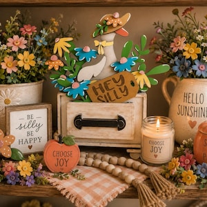 May include: A wooden goose cutout with a hat and flowers, a small drawer with a 'HEY SILLY' tag, a 'BE SILLY BE HAPPY' sign, a 'CHOOSE JOY' candle, a 'SWEET AS A PEACH' pumpkin jar, a 'CHOOSE JOY' apple, and a wooden bead garland on a checkered cloth.