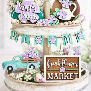 May include: A tiered wooden tray with floral decorations. The top tier has a purple flower with the words "happy bloom". The middle tier has a banner that says "bloom". The bottom tier has a turquoise truck and a sign that says "fresh flower market".