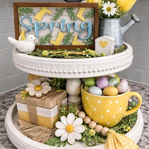 May include: A two-tiered white decorative tray with spring decorations. The top tier has a sign saying "Spring", a white bird, and a watering can with yellow tulips. The bottom tier has a yellow polka-dot cup filled with Easter eggs.
