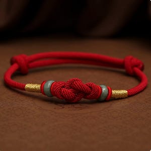 Puede incluir: Una pulsera de cuerda roja con un diseño de nudo central. La pulsera presenta detalles dorados y pequeñas cuentas verdes. El fondo es de color marrón cálido, resaltando el vibrante tono rojo de la pulsera.