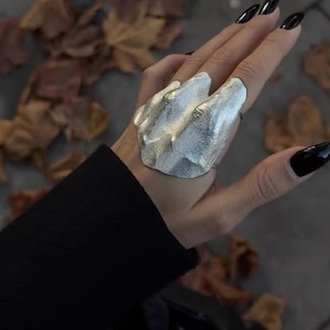 May include: A large, silver-toned statement ring featuring an irregular, textured surface with multiple raised, organic-shaped ridges. The ring covers a significant portion of the finger, showcasing a metallic finish with a hammered appearance.