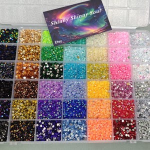 May include: A clear plastic organiser filled with a variety of colourful rhinestones. The rhinestones are arranged in a grid, showcasing shades of green, blue, pink, yellow, and more. A business card with the text "Shinny Shinny Soul" rests on top.