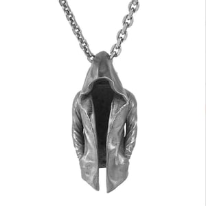 May include: Silver-toned pendant necklace featuring a hooded figure. The pendant has a textured finish, with a detailed hood and coat design. The necklace chain is a matching silver color, with a link chain style. The overall design is gothic and edgy.