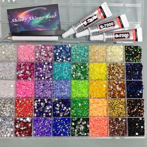 May include: A clear plastic organiser filled with colourful rhinestones in various shades. The organiser includes tubes of B-7000 glue, a business card that says "Shinny Shinny Soul", tweezers, and a pencil. The rhinestones are arranged in a grid.