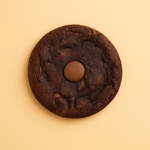 Op de afbeelding: Een grote, ronde, donkere chocoladekoek met een enkel, rond chocoladestuk in het midden. De koek is diepbruin van kleur en ligt op een lichtbeige achtergrond. De textuur van de koek lijkt enigszins ruw.