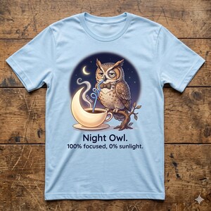 Puede incluir: Camiseta azul claro con un diseño de búho de dibujos animados. El búho, con pajarita, bebe de una taza de café en forma de media luna. El texto dice: "Night Owl. 100% focused, 0% sunlight".