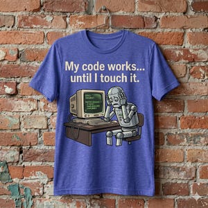 以下が含まれることがあります： 「My code works... until I touch it.」のテキストが入った青いTシャツ。コンピューターの前に座り、ストレスを感じている漫画のロボットが描かれています。コンピューターの画面にはコードが表示されています。