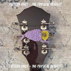Puede incluir: Primer plano de un clavijero de guitarra con clavijas de afinación. Una púa de guitarra amarilla se sujeta en un soporte de ganchillo lavanda. El texto "PATTERN ONLY - NO PHYSICAL PRODUCT" está en la parte superior e inferior de la imagen.