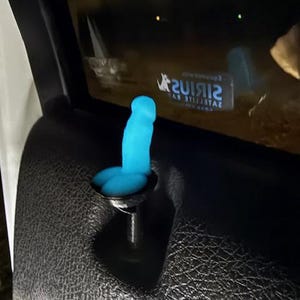 May include: A bright blue, novelty car door lock knob shaped like a phallus. The knob is installed in a black car door panel. The background is dark, with reflections visible in the car window.