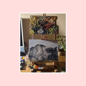 May include: A wooden easel holds a rectangular canvas featuring a grayscale painting of a curled-up cat. Above the canvas, a wooden palette is covered in thick, multi-colored oil paint. A single paintbrush rests on the bottom ledge of the easel.