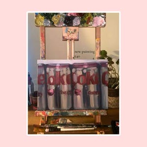 May include: A rectangular painting on a wooden easel featuring a series of vertical glass jars with red text that reads "pocky" and "cher" with small red circular accents. The painting is displayed on a wooden easel with paint brushes.