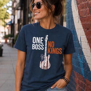 May include: A navy blue short-sleeve t-shirt featuring the white text ONE BOSS and orange text NO KINGS. Between the words is a graphic of an electric guitar with a distressed American flag pattern in red, white, and blue colors.