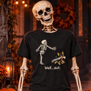 May include: A black short-sleeved t-shirt featuring a graphic of a skeleton reaching towards a small dog holding a bone in its mouth. Below the graphic, the text reads "Well...shit." The design is printed in white, yellow, and black ink.