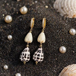 May include: A pair of gold hoop earrings with white and black seashell pendants. The earrings are adorned with small clear stones and hang from a gold hoop. The background features pearls and seashells on a dark surface.