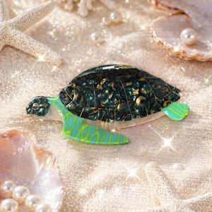 May include: A decorative sea turtle brooch with a dark green and gold shell, and light green flippers with yellow accents. The brooch is set on a sandy surface with seashells, pearls, and starfish.