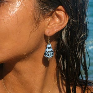 Puede incluir: Primer plano de un pendiente dorado con un colgante estampado en azul y negro. El pendiente cuelga de una oreja, con el pelo mojado y el mar al fondo. El colgante tiene forma de lágrima.