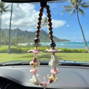 Puede incluir: Un accesorio para coche con un diseño tipo lei. La guirnalda está compuesta por cuentas de madera, detalles de conchas marinas y delicados colgantes de flores de colores claros. El fondo muestra una vista panorámica de un paisaje tropical con palmeras y el océano.