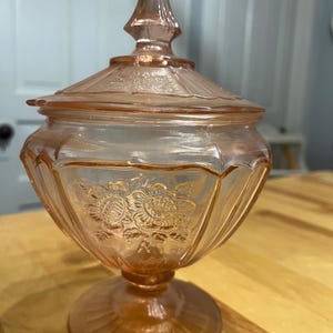 Pode incluir: Um pote de vidro rosa claro para doces com tampa correspondente apresentando um remate pontiagudo. A ta&ccedil;a tem uma base de pedestal e &eacute; decorada com padr&otilde;es florais em relevo e sulcos verticais ao redor do corpo e da tampa.