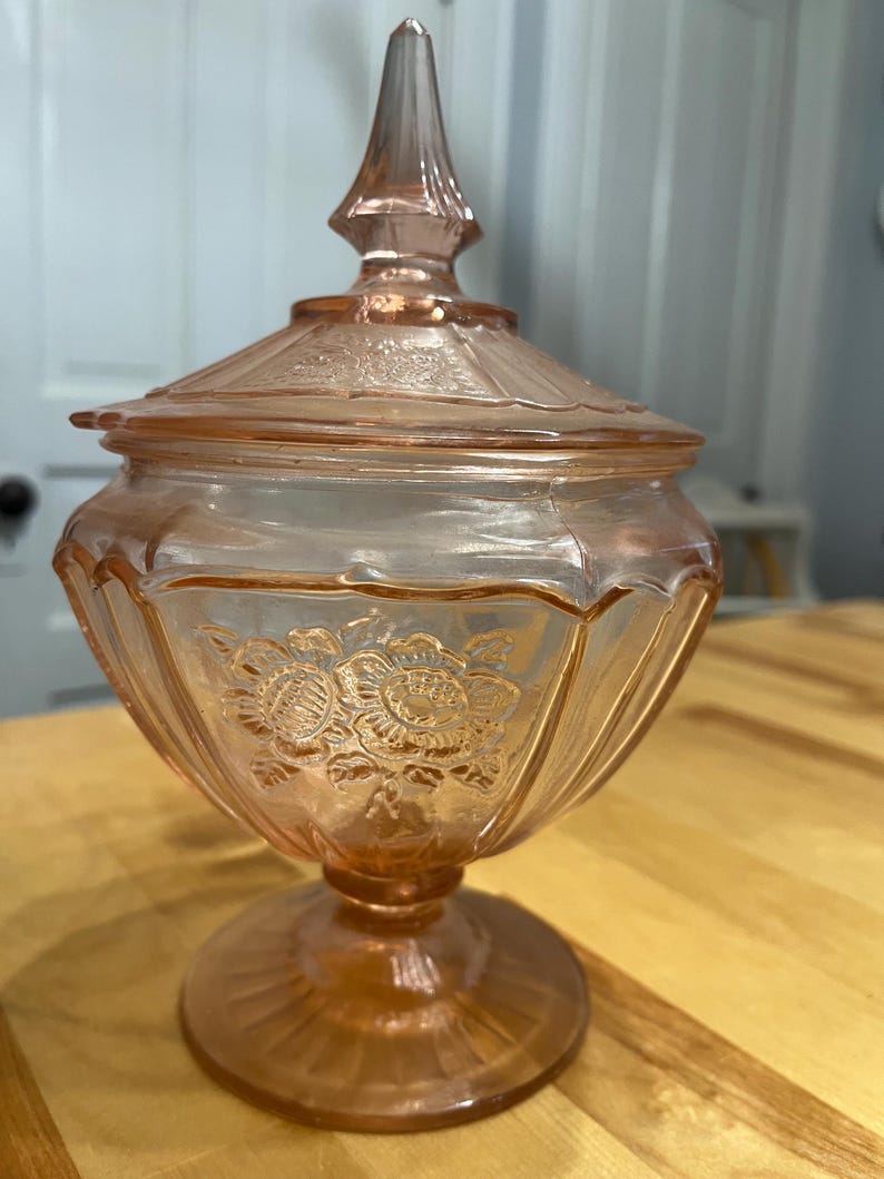 Pode incluir: Um pote de vidro rosa claro para doces com tampa correspondente apresentando um remate pontiagudo. A ta&ccedil;a tem uma base de pedestal e &eacute; decorada com padr&otilde;es florais em relevo e sulcos verticais ao redor do corpo e da tampa.